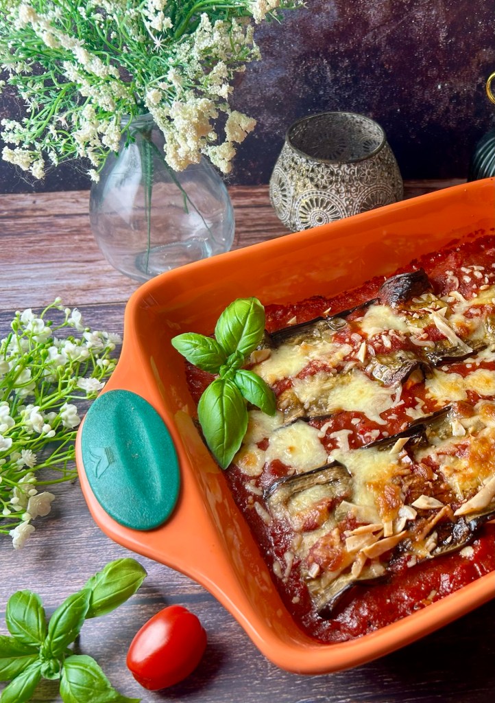 Ein Gericht mit Auberginen-Ricotta-Röllchen in einer orangen Auflaufform, überzogen mit Käse und Tomatensauce, garniert mit frischem Basilikum und umgeben von Kräutern und Tomaten.