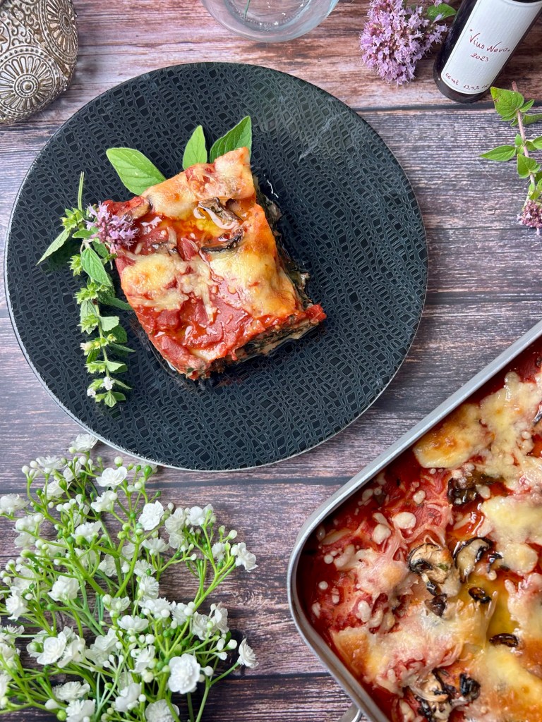 Ein Teller mit einer Portion Spinatlasagne, garniert mit frischen Kräutern, auf einem schwarzen Teller vor einem Backblech mit mehr Lasagne.