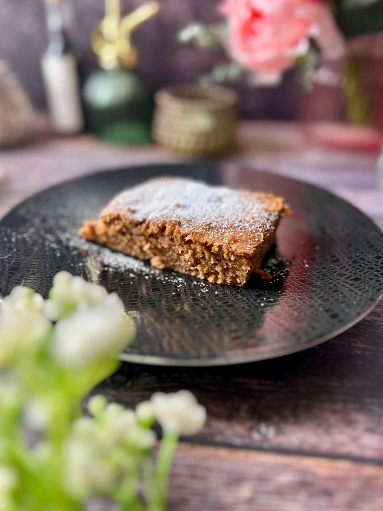 Ein Stück Apfelkuchen auf einem dekorativen schwarzen Teller, mit Puderzucker bestäubt, umgeben von unscharfen Blüten und dekorativen Objekten im Hintergrund.