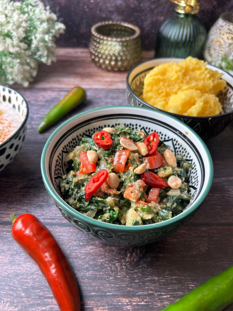 Eine porzellanartige Schüssel mit Ifisashi, einem sambischen Erdnuss-Gemüse-Eintopf, garniert mit roten Chilischoten und Erdnüssen. Im Hintergrund sind grüne Chilis, Nshima (Maisbrei) und weitere dekorative Elemente zu sehen.