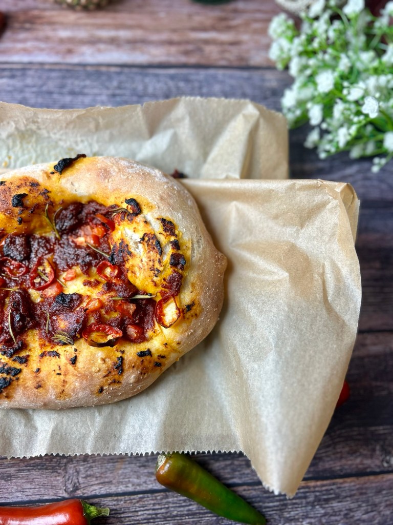 Ein frisch gebackenes Fladenbrot mit einer würzigen Tomaten-Curry-Paste, garniert mit Chili und Rosmarin, auf Backpapier präsentiert.